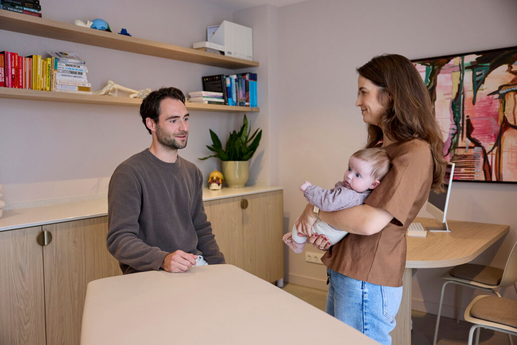 Osteopaat in gesprek met een moeder die haar baby in haar armen houdt.