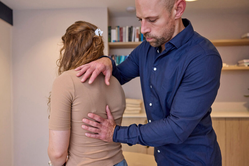 Osteopaat onderzoekt de rug van een zittende vrouw met zijn handen op haar schouder en rug.
